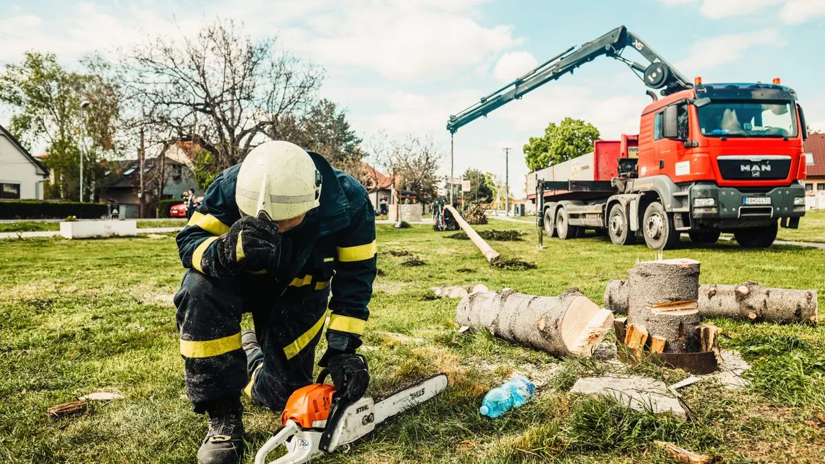 Arborist in safety gear using chainsaw beside truck with crane in an outdoor setting.